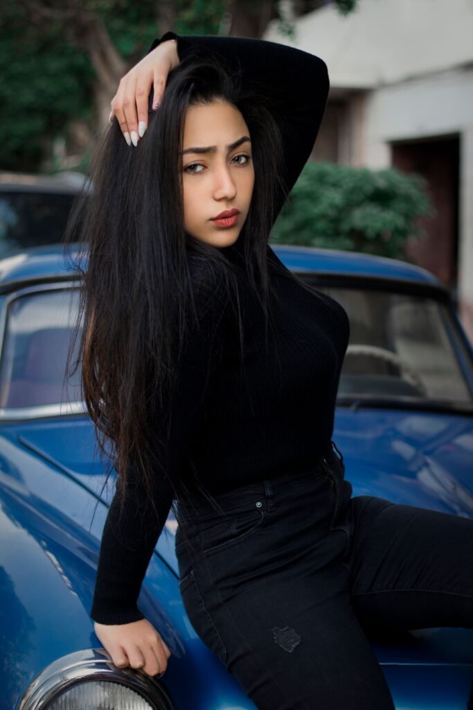 Stylish young woman with long hair poses confidently on a vintage blue car outdoors.